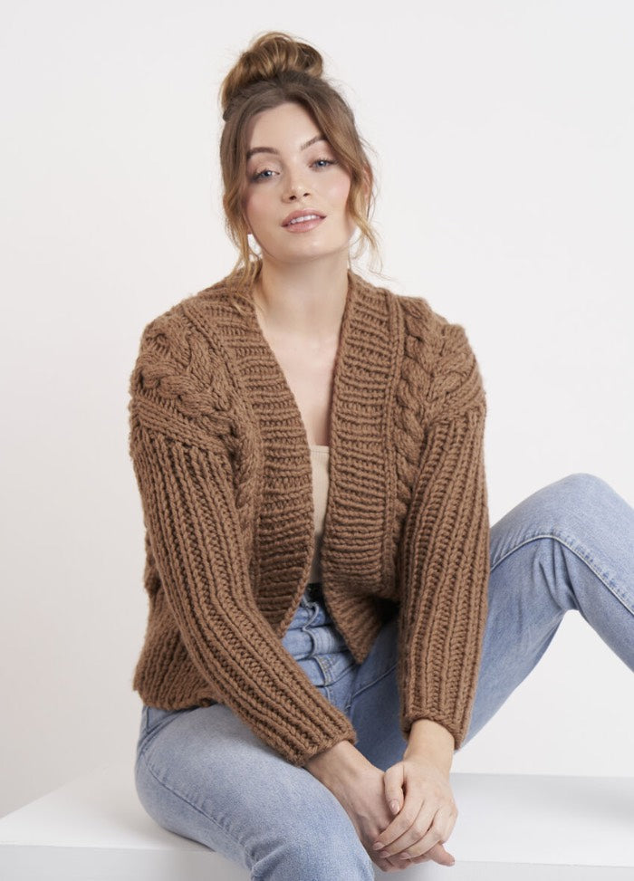 A person sits casually on a white surface, wearing a cozy brown knitted sweater and light blue jeans. Their hair is in a loose bun, conveying a relaxed and comfortable vibe.