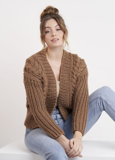 A person sits casually on a white surface, wearing a cozy brown knitted sweater and light blue jeans. Their hair is in a loose bun, conveying a relaxed and comfortable vibe.