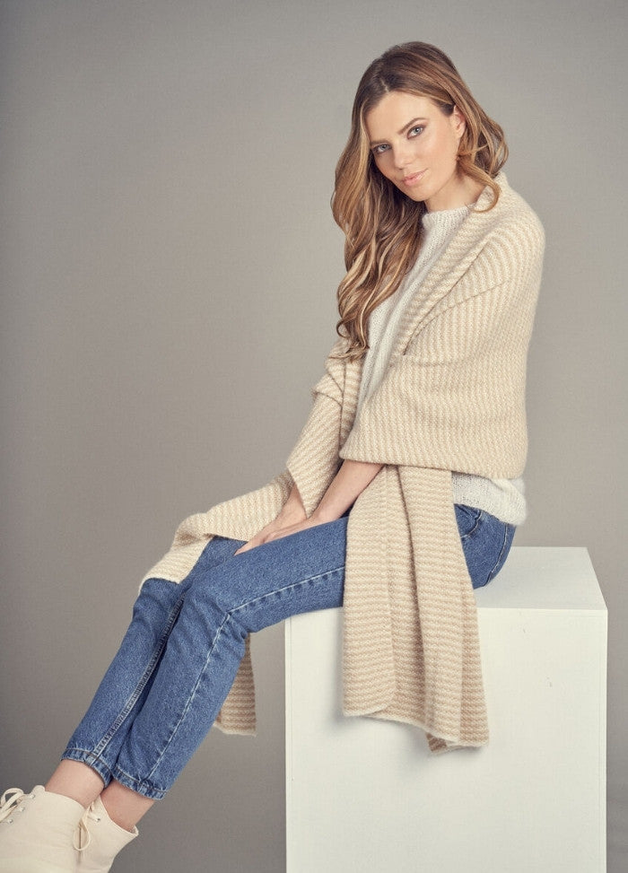 A woman with long hair sits on a white cube, draped in a beige striped shawl over a light sweater and blue jeans. She has a serene expression, against a plain gray background.
