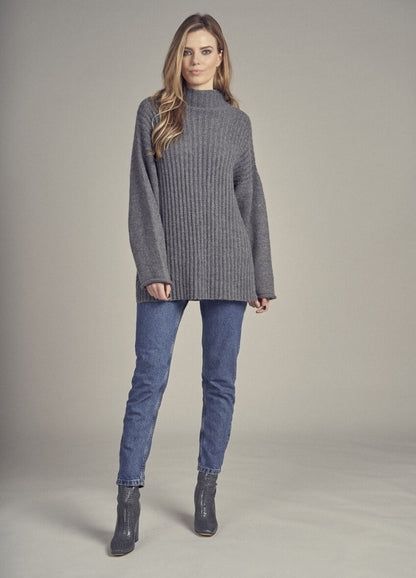 A woman stands confidently in a gray ribbed sweater and blue jeans against a neutral backdrop. She wears dark ankle boots, conveying a relaxed, stylish look.