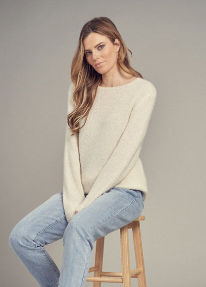 A person with long hair, wearing a cream sweater and light blue jeans, sits on a wooden stool. The background is neutral, conveying a relaxed mood.