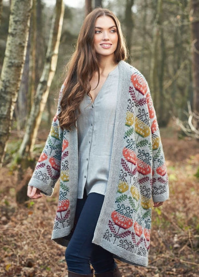 A woman in a colorful, floral-patterned cardigan and gray shirt smiles while walking through a serene forest. The background features tall trees and autumn leaves.