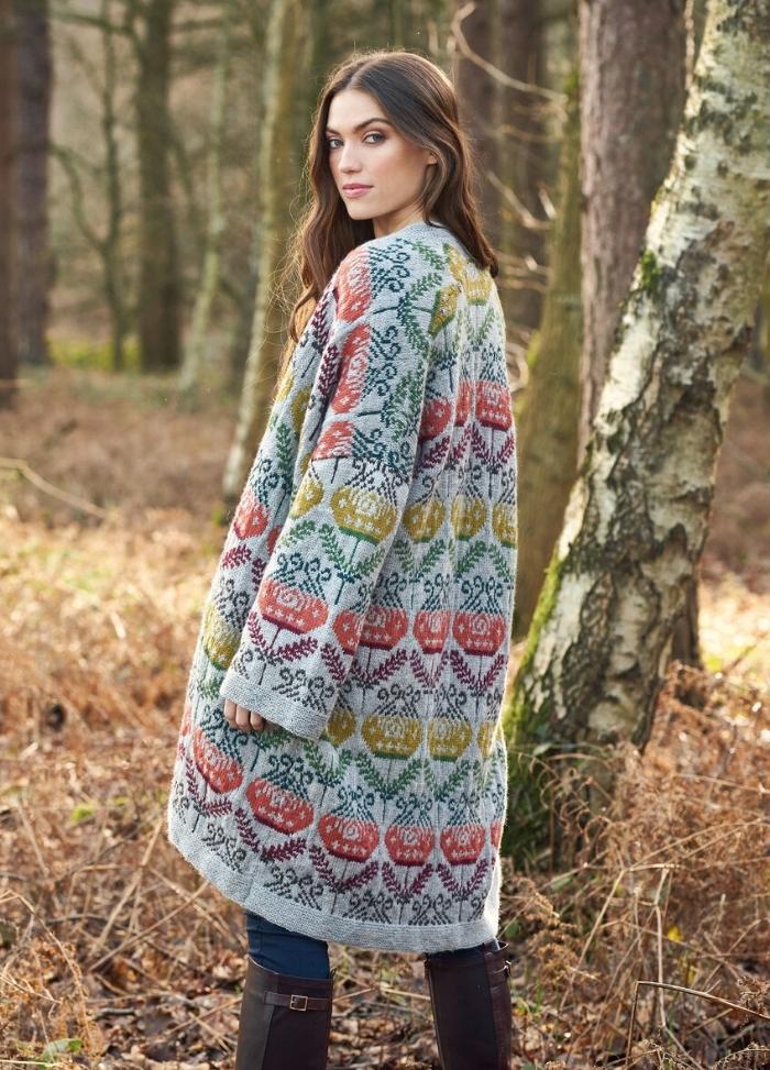 A woman in a colorful, floral-patterned cardigan and gray shirt smiles while walking through a serene forest. The background features tall trees and autumn leaves.