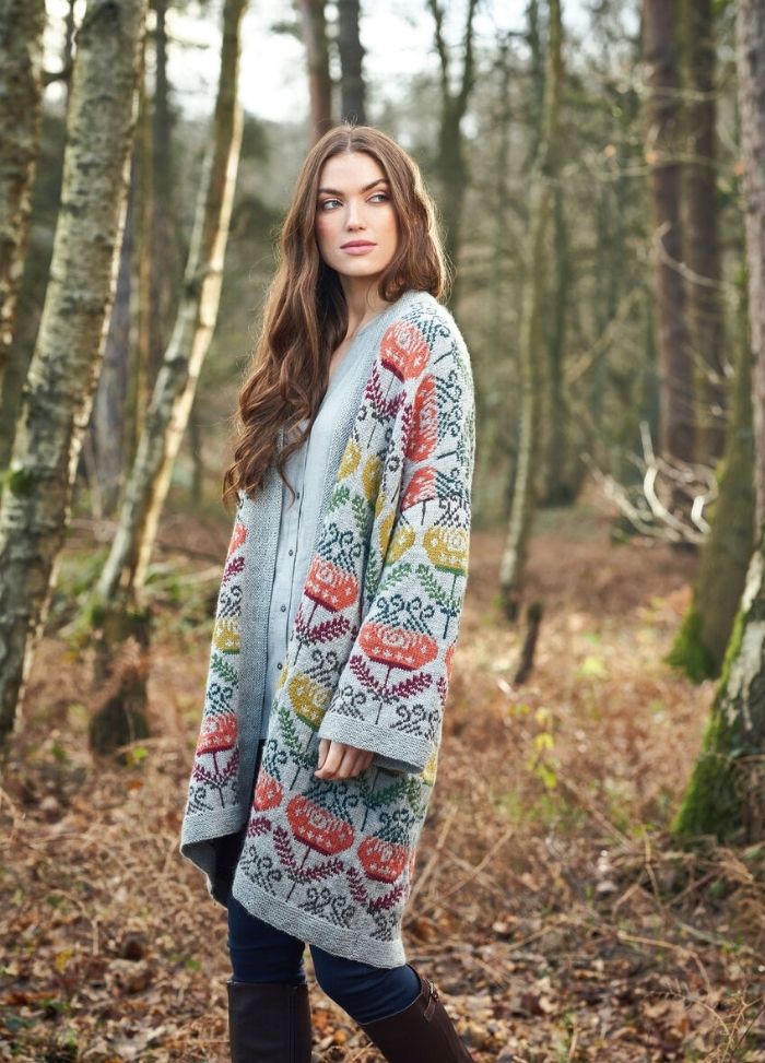 A woman with long hair stands in a forest, wearing a colorful, patterned cardigan and blue jeans. The scene is calm, with autumn leaves on the ground.