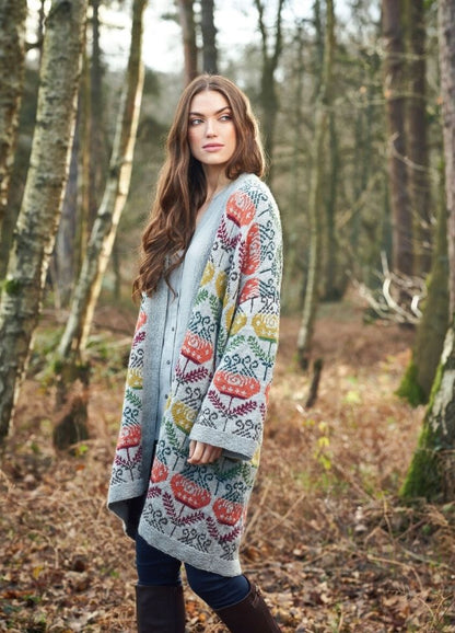 A woman with long hair stands in a forest, wearing a colorful, patterned cardigan and blue jeans. The scene is calm, with autumn leaves on the ground.