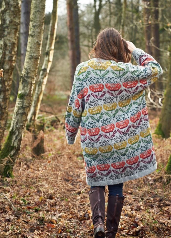 A woman walks away in a forest, wearing a colorful, patterned coat with floral designs. The scene feels peaceful and autumnal, with scattered leaves.