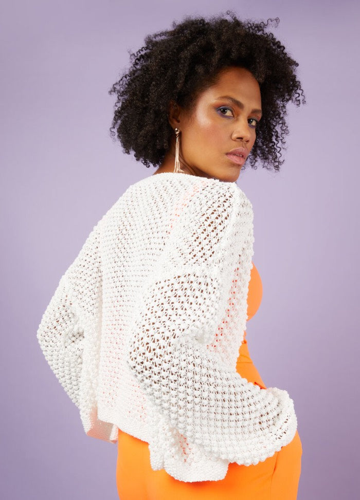Woman with curly hair wearing a white crochet sweater over an orange dress stands against a purple background, conveying a confident and stylish mood.