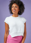 Smiling woman with curly hair wears a white sleeveless knit top and pink pants against a lavender background, conveying a cheerful, stylish vibe.
