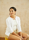 A woman in a white sweater and shorts sits on a yellow chair against a beige background. She looks to the side with a relaxed expression, wearing large hoop earrings.