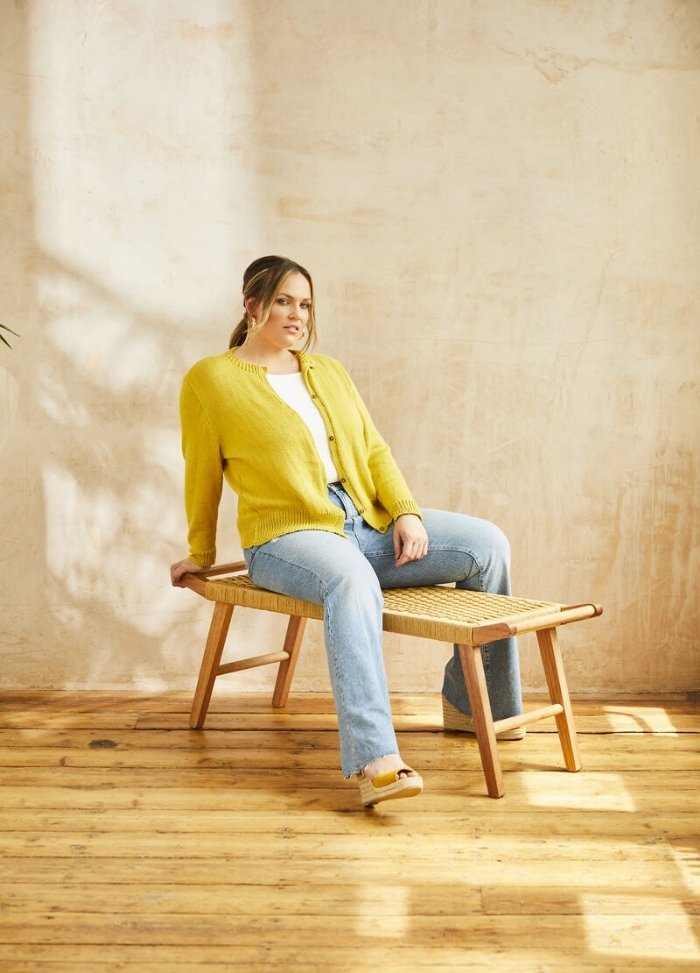 A person wearing a yellow cardigan and jeans sits on a woven bench in a sunlit room with wooden floors and textured beige walls, exuding a relaxed vibe.