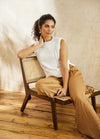 Woman in a white sleeveless top and tan pants sits stylishly on a wooden chair. She gazes to the side, exuding a relaxed and thoughtful mood.