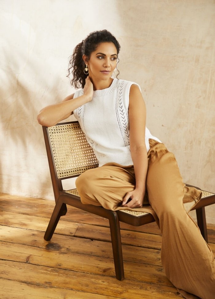 Woman in a white sleeveless top and tan pants sits stylishly on a wooden chair. She gazes to the side, exuding a relaxed and thoughtful mood.