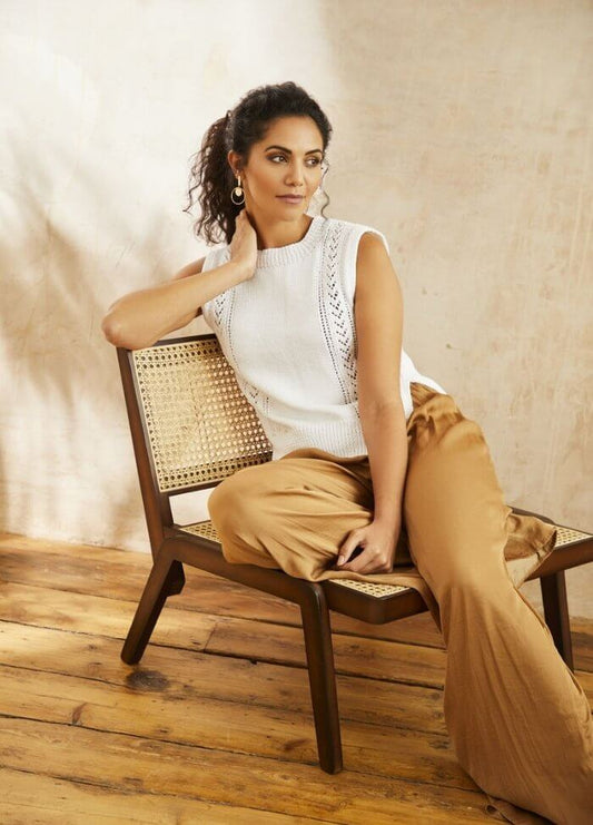 Woman in a white sleeveless top and tan pants sits stylishly on a wooden chair. She gazes to the side, exuding a relaxed and thoughtful mood.