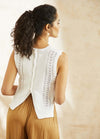 Woman with curly ponytail wearing a white knit sleeveless top, featuring intricate patterns and back buttons, paired with tan pants, standing in soft light.
