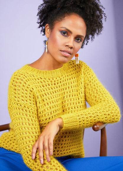 Woman wearing a bright yellow knit sweater and blue pants, sitting on a wooden chair with a soft purple backdrop. Her expression is calm and confident.