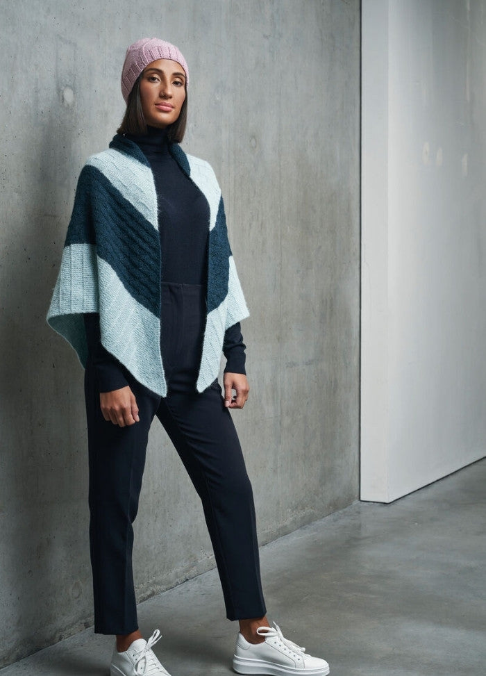 A woman in a pink knit hat and colorful shawl stands confidently against a concrete wall. She wears a black outfit with white sneakers, exuding a modern, chic vibe.