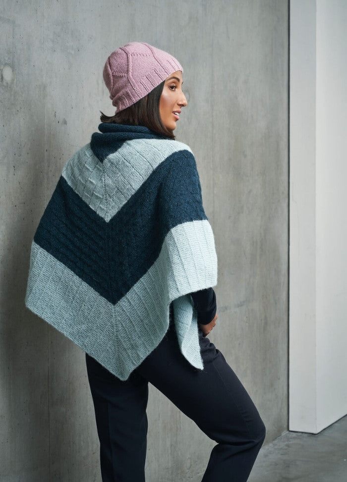 Woman in pink knit hat and blue, black, and white chevron-patterned poncho stands against a gray concrete wall, expressing a calm demeanor.