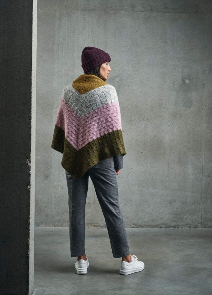 A person in a colorful knit shawl stands against a gray concrete wall. They're wearing a maroon beanie, gray pants, and white sneakers, conveying a cozy, casual vibe.