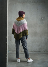 A person in a colorful knit shawl stands against a gray concrete wall. They're wearing a maroon beanie, gray pants, and white sneakers, conveying a cozy, casual vibe.