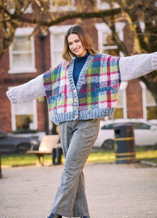 Woman smiling and posing outdoors with arms outstretched. She wears a colorful plaid cardigan and gray pants in a park setting. The mood is cheerful.