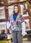 A woman smiles outdoors wearing a colorful plaid cardigan with large buttons, paired with a navy turtleneck and gray pants. The background is a blurred building and trees.
