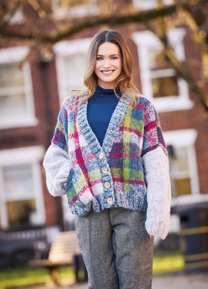 A woman smiles outdoors wearing a colorful plaid cardigan with large buttons, paired with a navy turtleneck and gray pants. The background is a blurred building and trees.