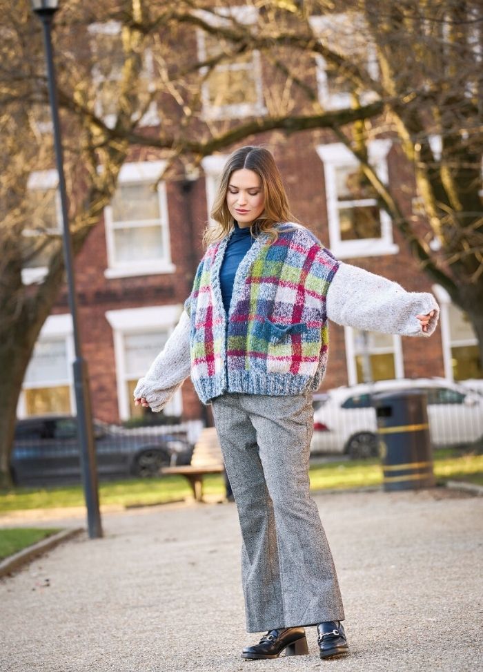 A woman in a colorful, plaid cardigan and gray trousers walks in a park with autumn trees. She appears relaxed, exuding a casual and calm demeanor.