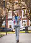 A woman walks confidently in a park. She wears a colorful checkered cardigan, blue turtleneck, and gray pants. Trees and a brick building are in the background.
