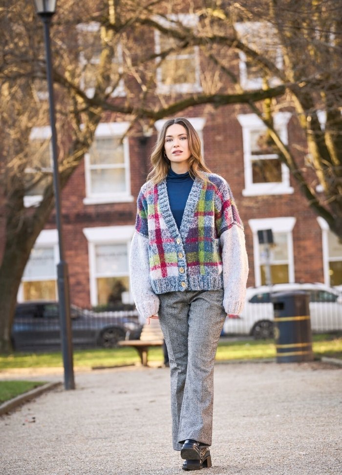 A woman walks confidently in a park. She wears a colorful checkered cardigan, blue turtleneck, and gray pants. Trees and a brick building are in the background.