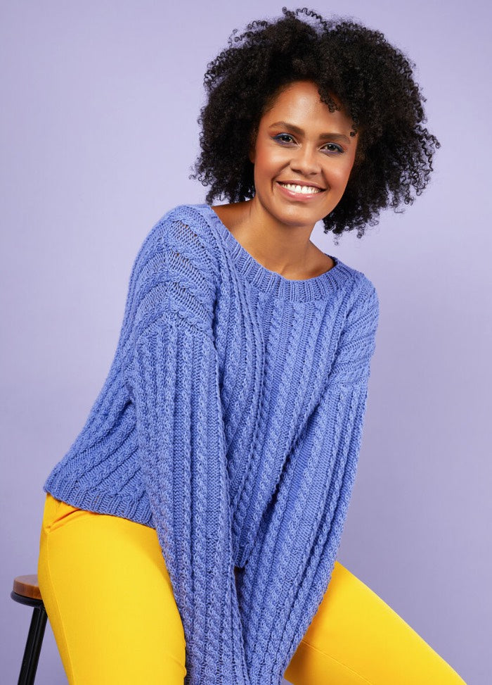Smiling person with curly hair wearing a blue textured sweater and bright yellow pants, sitting on a stool against a lavender background.