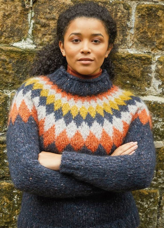 Person with curly hair, wearing a patterned, colorful sweater, stands confidently with arms crossed against a textured stone wall. The tone is warm and assertive.