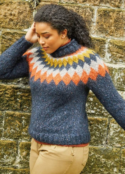 Woman in a cozy, patterned sweater with blue, orange, and yellow accents leans thoughtfully against a textured stone wall, exuding warmth and contemplation.