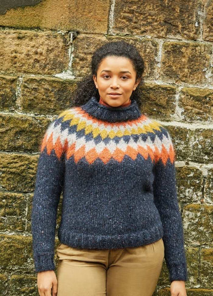 Woman in a blue knitted sweater with colorful geometric patterns, stands against a textured stone wall. She has a calm, relaxed expression.