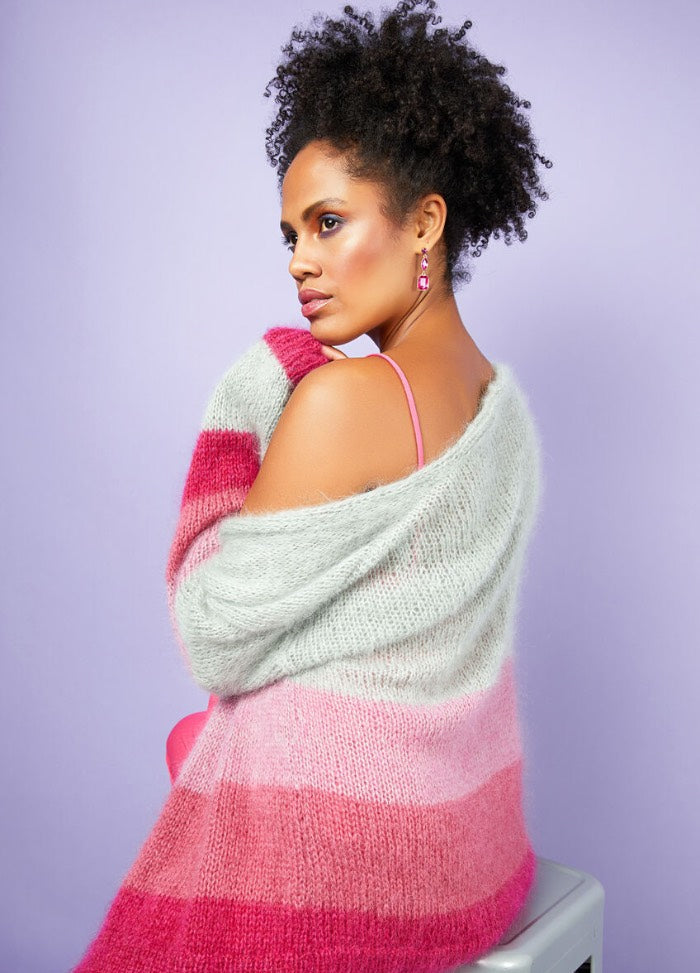 Woman with curly hair in a pink-and-white striped sweater sits against a pastel purple background, exuding confidence and elegance.