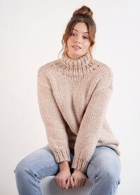 A person with a relaxed expression sits against a white background, wearing a chunky beige turtleneck sweater and light blue jeans, conveying a cozy and casual vibe.