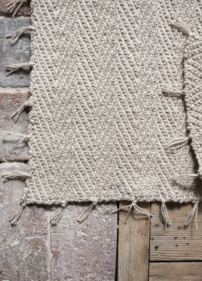 Close-up of a textured beige woven rug with chevron patterns and fringed edges, laid over a weathered brick and wooden floor, creating a rustic feel.