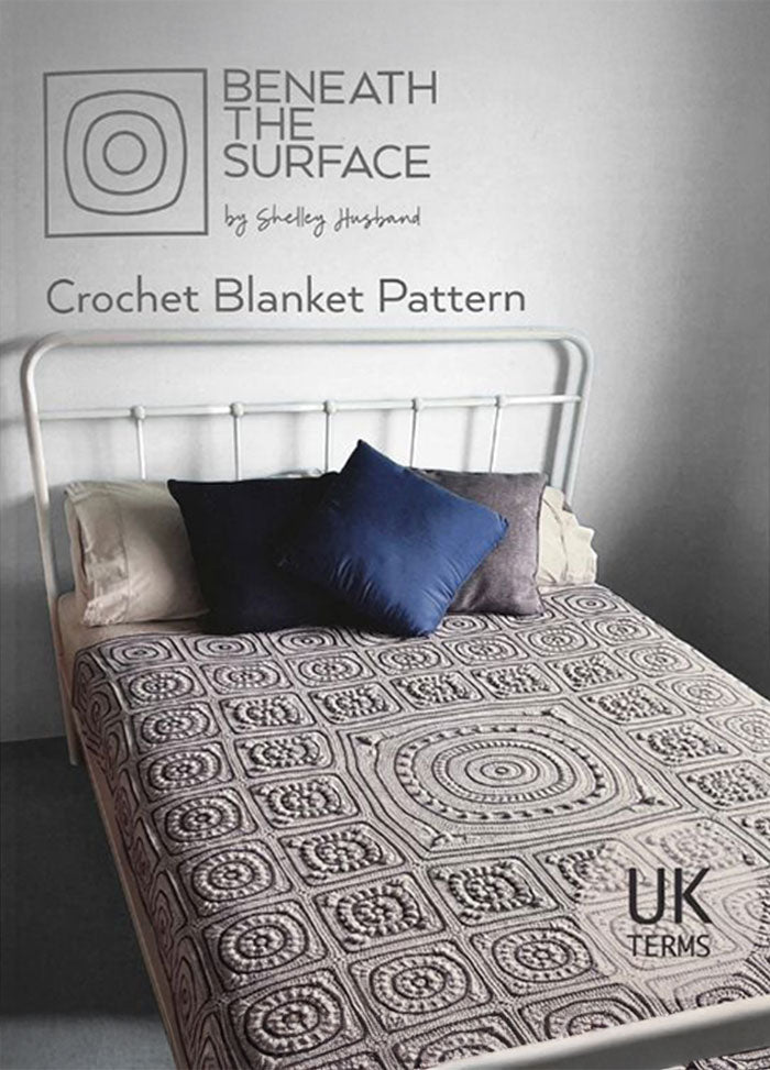 A metal-framed bed features a detailed crochet blanket with circular and square patterns. Two pillows, one dark blue, add contrast. Text reads "Beneath the Surface: Crochet Blanket Pattern by Shelley Husband" in UK terms. The scene feels cozy and inviting.