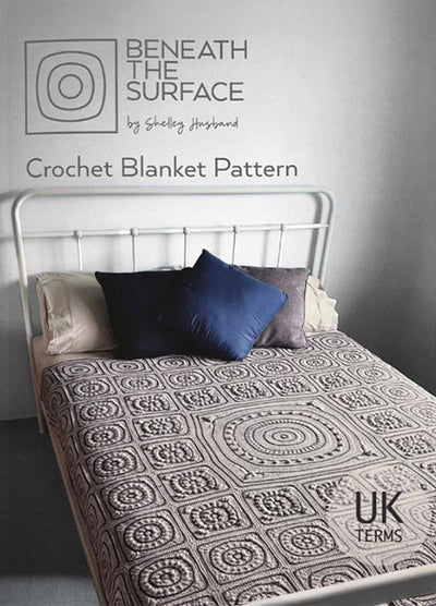 A metal-framed bed features a detailed crochet blanket with circular and square patterns. Two pillows, one dark blue, add contrast. Text reads "Beneath the Surface: Crochet Blanket Pattern by Shelley Husband" in UK terms. The scene feels cozy and inviting.