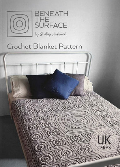 A metal-framed bed features a detailed crochet blanket with circular and square patterns. Two pillows, one dark blue, add contrast. Text reads "Beneath the Surface: Crochet Blanket Pattern by Shelley Husband" in UK terms. The scene feels cozy and inviting.