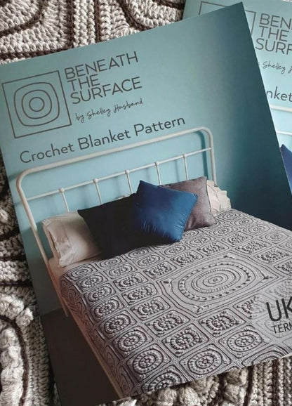 Crochet blanket pattern booklet titled "Beneath the Surface" shows a geometric-patterned blanket on a white bed with blue pillows, evoking a cozy vibe.