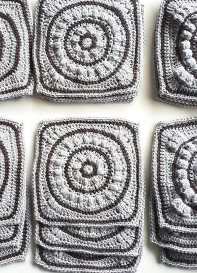 Gray crochet squares with intricate circular patterns are neatly stacked in rows, showcasing the textured craftsmanship and creating a sense of orderly repetition.