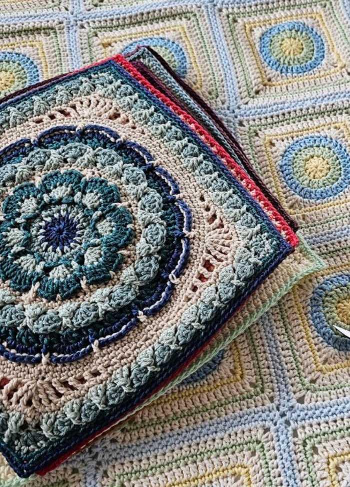Colorful crochet pillow on a patterned afghan, featuring intricate floral and geometric designs in shades of blue, green, and beige. Warm and cozy feel.