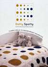 Crochet book cover titled "Dotty Spotty" by Shelley Husband, features a blanket with mustard and navy circles on squares. Plush sheep toys sit on the bed, conveying warmth and coziness.
