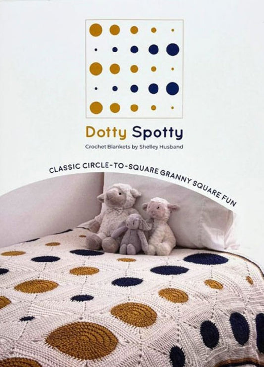 Crochet book cover titled "Dotty Spotty" by Shelley Husband, features a blanket with mustard and navy circles on squares. Plush sheep toys sit on the bed, conveying warmth and coziness.
