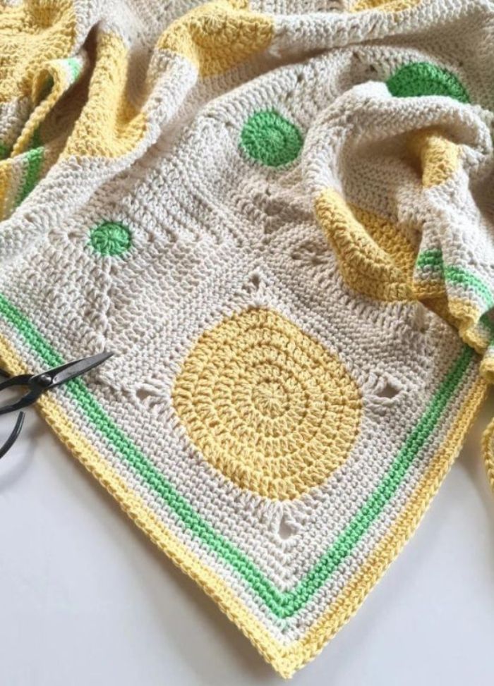 Intricately crocheted blanket with a beige base, featuring yellow and green circular patterns, and a green-bordered edge. Scissors rest on it.