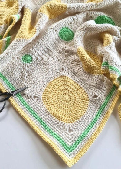 Intricately crocheted blanket with a beige base, featuring yellow and green circular patterns, and a green-bordered edge. Scissors rest on it.