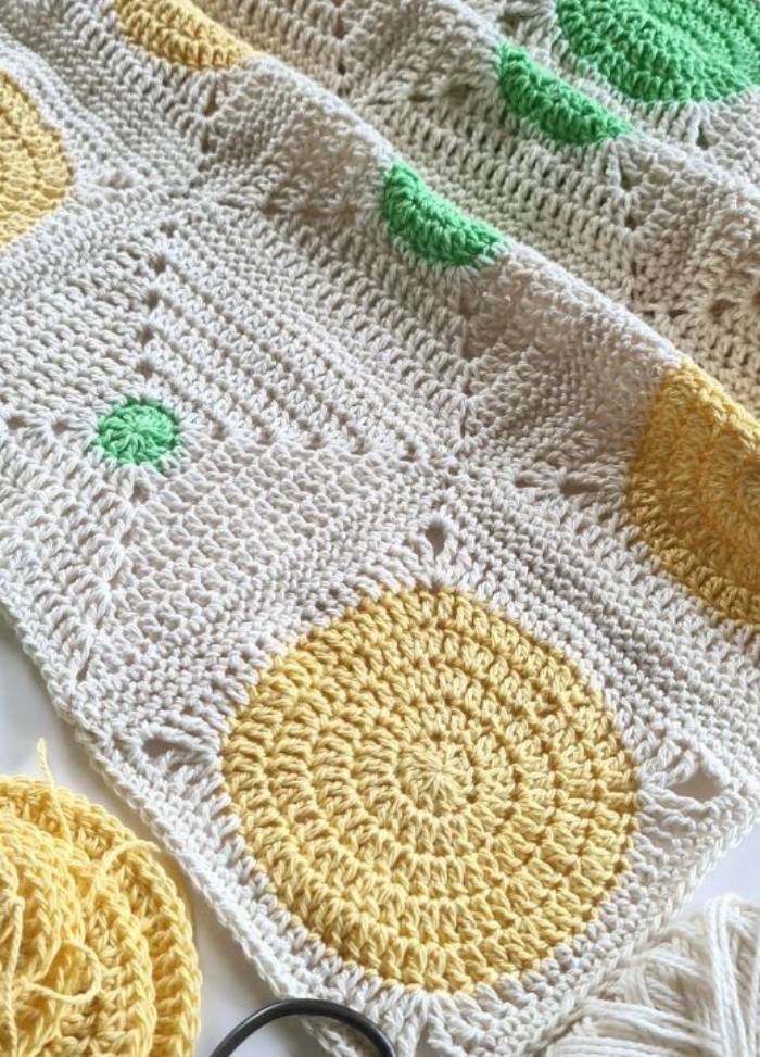 Crocheted blanket with circular patterns in yellow and green on a cream background. Yarn balls are visible, suggesting a warm, creative atmosphere.