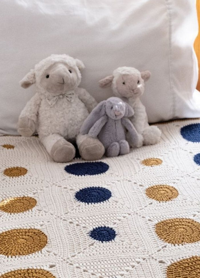 Three plush toys—a gray bunny and two sheep—sit on a bed with a white, crochet blanket featuring navy and gold polka dots, evoking a cozy atmosphere.
