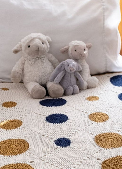 Three plush toys—a gray bunny and two sheep—sit on a bed with a white, crochet blanket featuring navy and gold polka dots, evoking a cozy atmosphere.