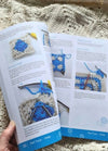 Open crochet instruction book on a textured knitted blanket. Pages show step-by-step guides with images of blue and white crochet squares.